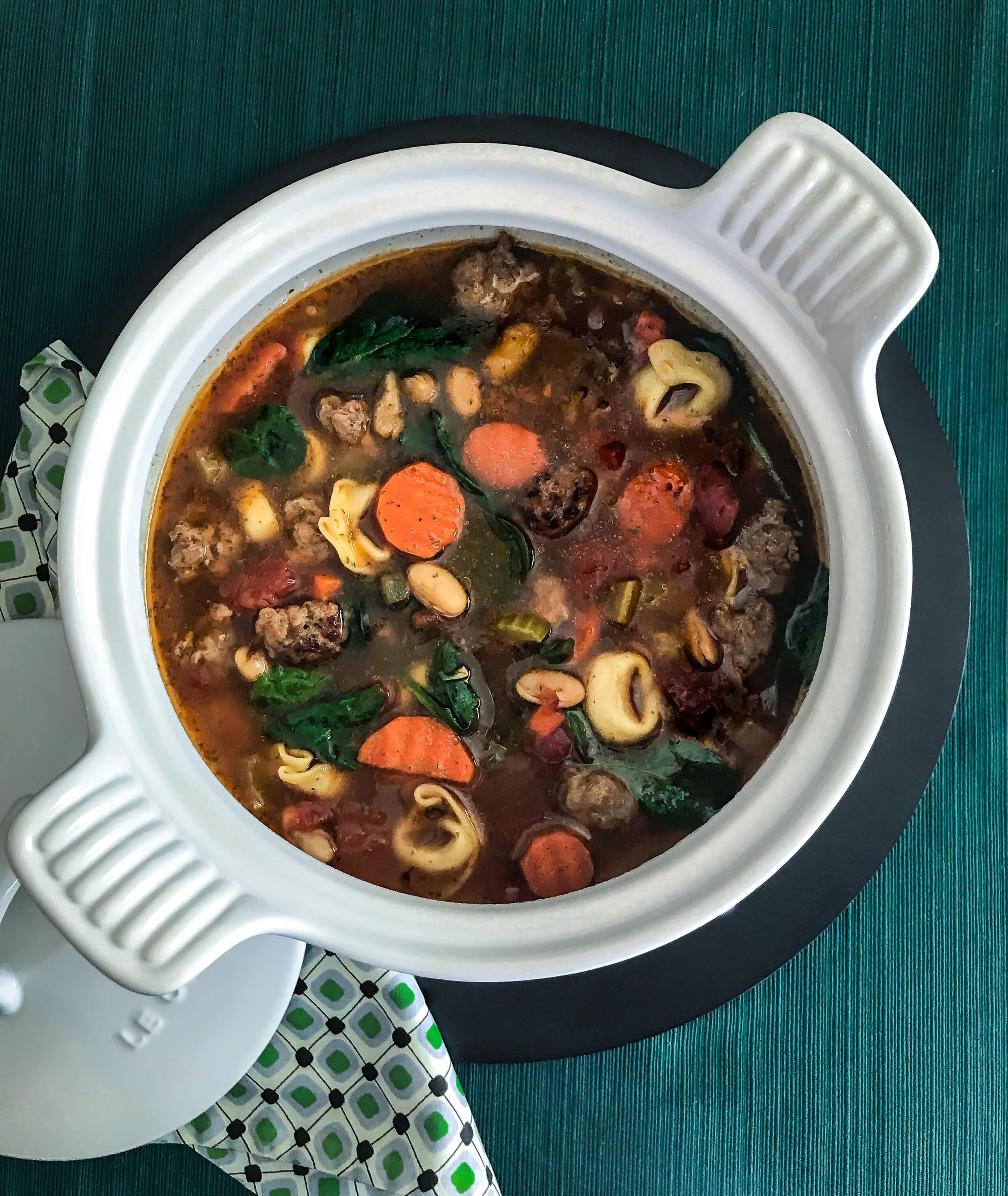 A white soup tureen filled with Italian Sausage and Bean Soup on a green background.