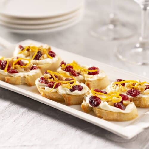 A white platter filled with cranberry orange crostini on a marble counter. Wine glasses and appetizer plates are in the background.