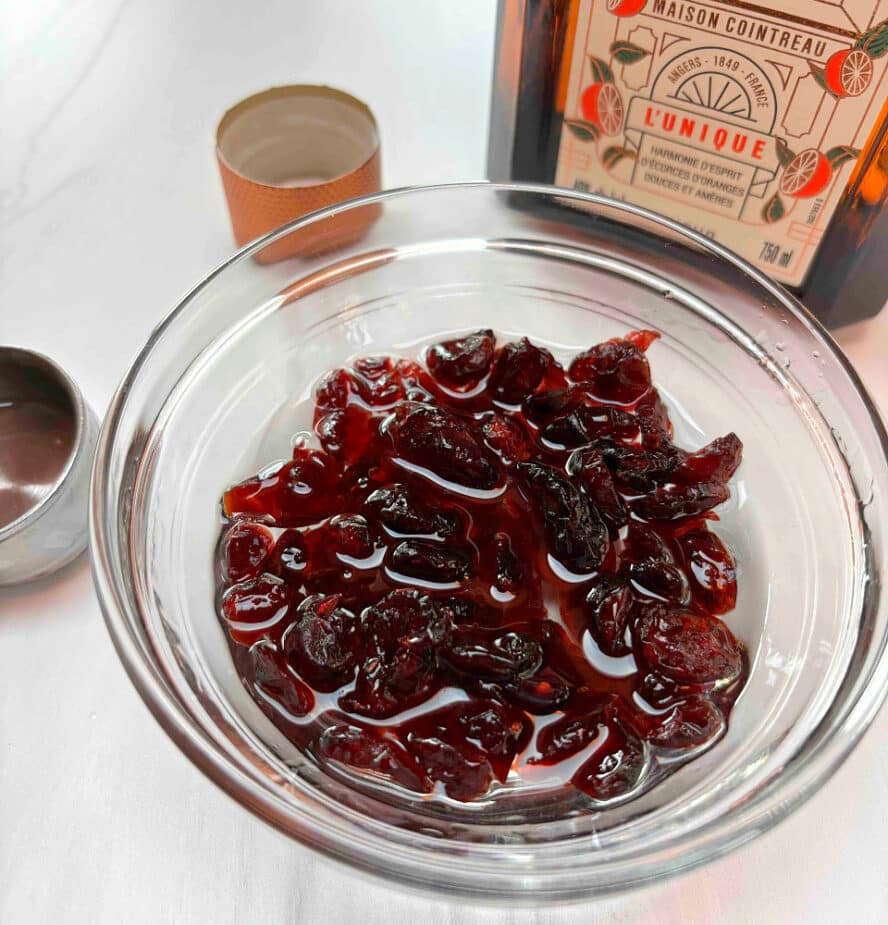 A small glass bowl filled with dried cranberries covered with Cointreau liqueur. A bottle of Cointreau is in the background.