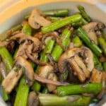A white bowl filled with sauteed asparagus, mushrooms and red onions