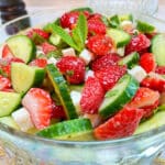 A glass bowl filled with diced strawberries, cucumbers, and feta cheese on a white table and rattan placemat. A dark wood pepper grinder is in the background