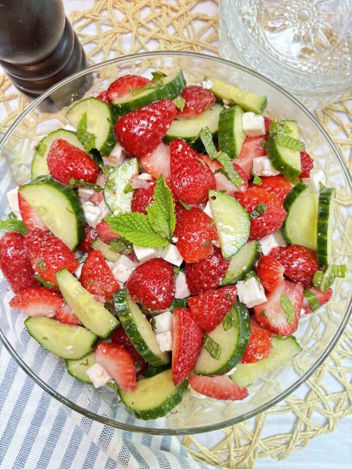 A glass bowl filled with diced strawberries, cucumbers, and feta cheese on a white table and rattan placemat. A dark wood pepper grinder is on the side.