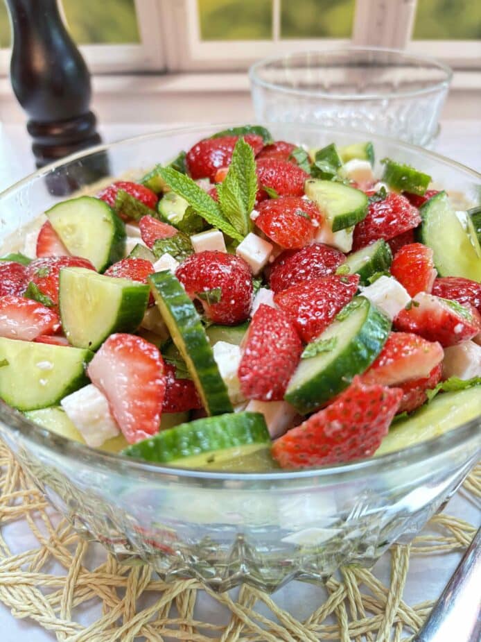 A glass bowl filled with bright red cut strawberrries, sliced cucumbers and diced feta cheese. It sits on a white table with a rattan placemat. Glass serving bowls and a wooden pepper grinder are in the background.