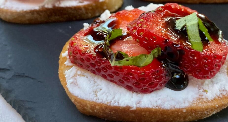 A thin crostini topped with a thin layer of goat cheese, 2 strawberry slices and a drizzle of balsamic vinegar glaze and sliced basil sitting on a slate tray.