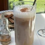 A glass of iced cinnamon coffee on a white table with cinnamon sticks and cinnamon syrup in the background