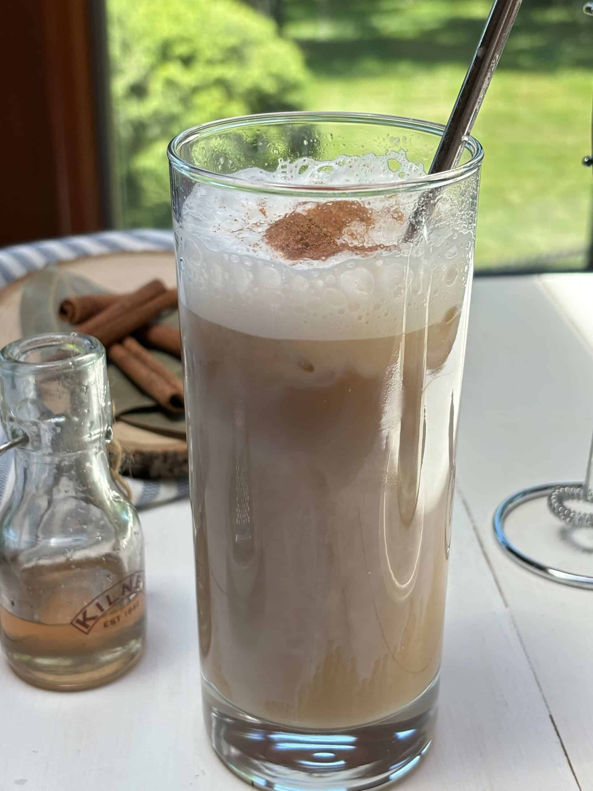 A glass of iced cinnamon coffee on a white table with cinnamon sticks and cinnamon syrup in the background