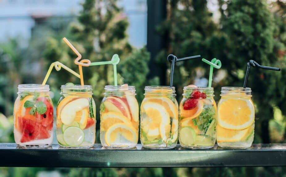6 mason jars filled with slices of fruit, herbs and water on a black iron table outside. Each glass has a fun colored straw to drink out of.