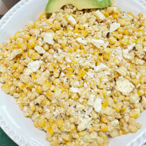 Corn salad in a white decorative serving bowl garnished with avocados on a green check tablecloth