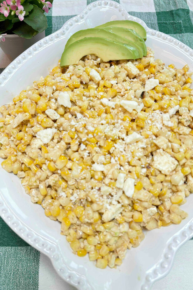 Corn salad in a white decorative serving bowl garnished with avocados on a green check tablecloth