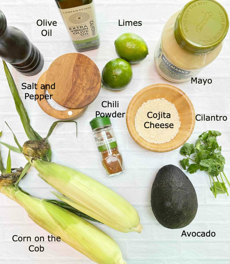 Ingredients used to make a street corn salad on a white tile table. They include corn on the cob, avocado, cilantro, crumbled cojita cheese, chili powder, mayonnaise, limes, olive oil and salt and pepper