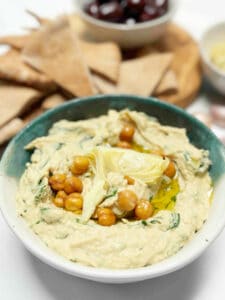 A green and white ceramic bowl filled with spinach and artichoke hummus with pita bread wedges in the background