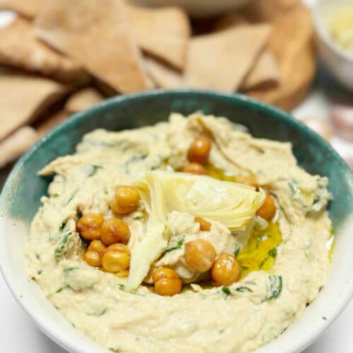 A green and white ceramic bowl filled with spinach and artichoke hummus with pita bread wedges in the background