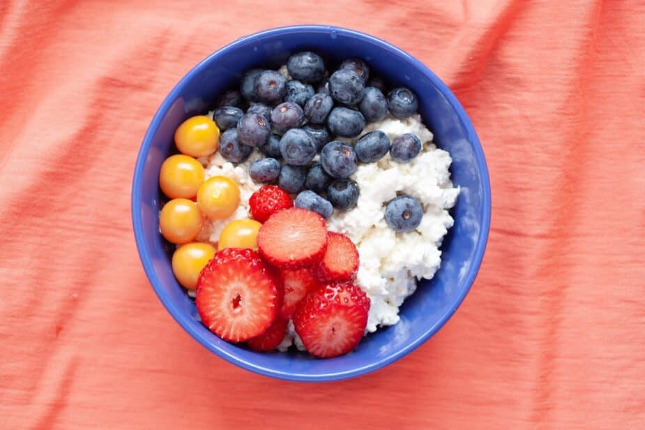 A blue bowl filled with cottage cheese and topped with blueberries, strawberries and gooseberries. It is sitting on a peach colored tablecloth