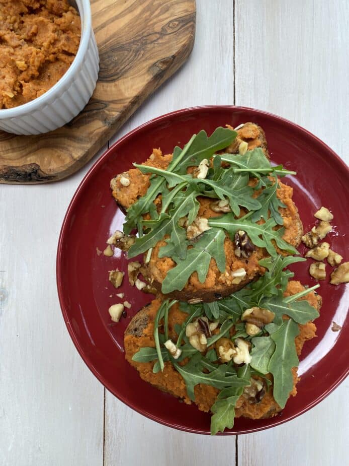 A red plate with whole grain bread spread with sundried tomato bean dip topped iwht arugula and chopped walnuts