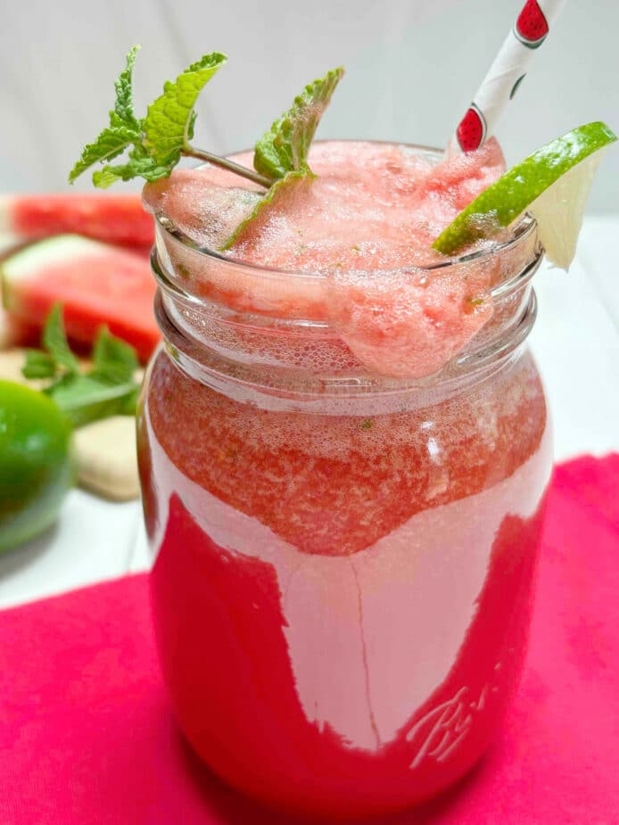 Watermelon Cooler in a glass with a wedge of lime and sprig of mint. Sitting on a red placemat on a white picnic table.