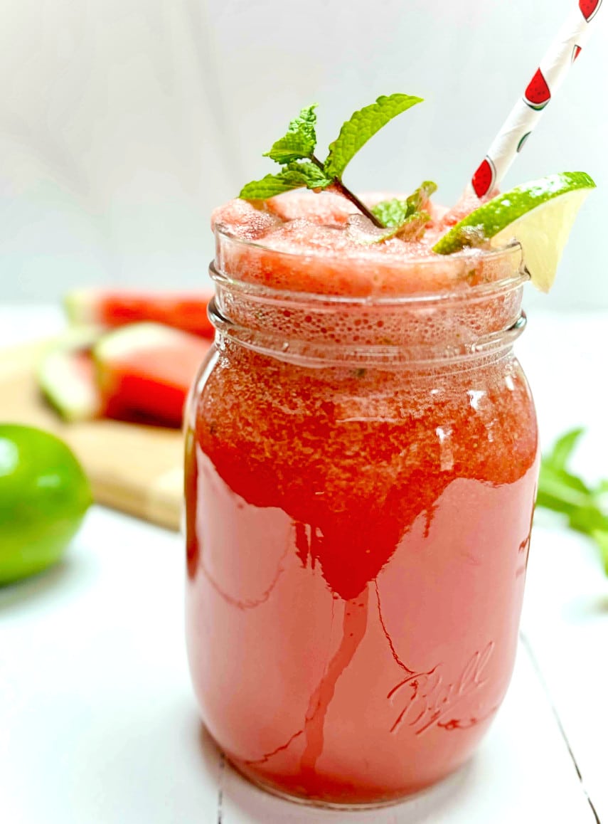 Watermelon lime cooler in a clear glass ball jar with a wedge of lime and mint sprig. It is sitting on a white picnic table.