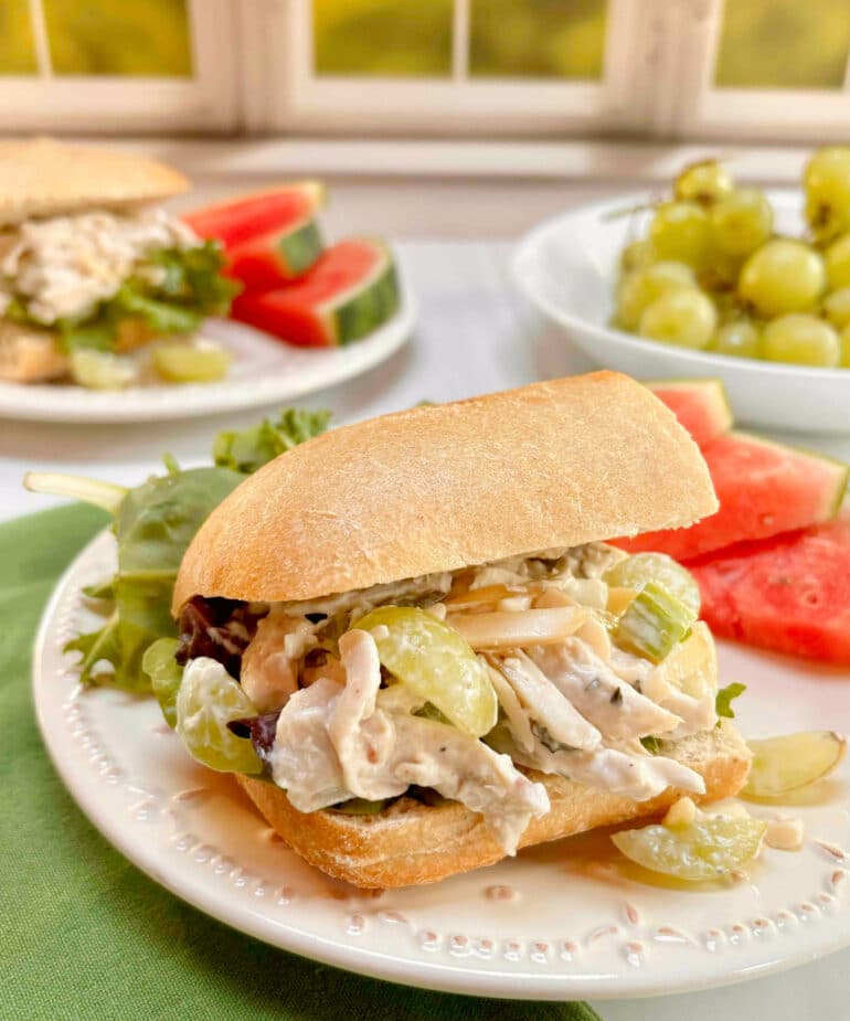 Chicken salad with grapes and almonds on a ciabatta roll with a green salad and sliced watermelon in the background