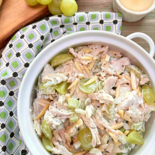 A white bowl filled with chicken salad with grapes and almonds on a picnic table. A green, white and black napkin surrounds the bowl with grapes and a roll in the background.