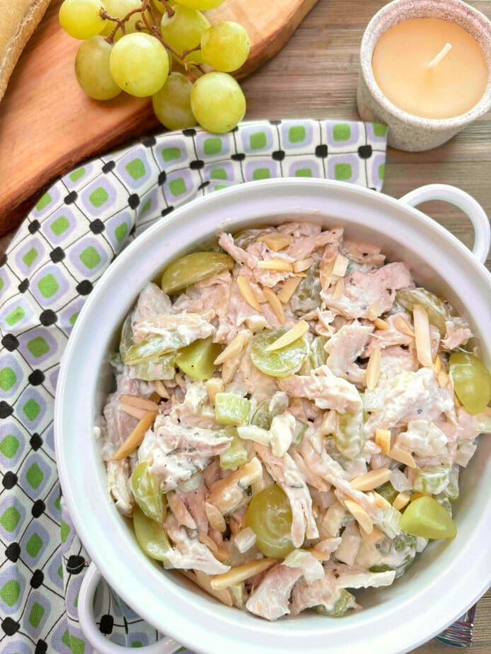 A white bowl filled with chicken salad with grapes and almonds on a picnic table. A green, white and black napkin surrounds the bowl with grapes and a roll in the background.