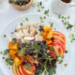 A white bowl filled with roasted chicken, butternut squash, apples and topped with microgreens on a marble counter.