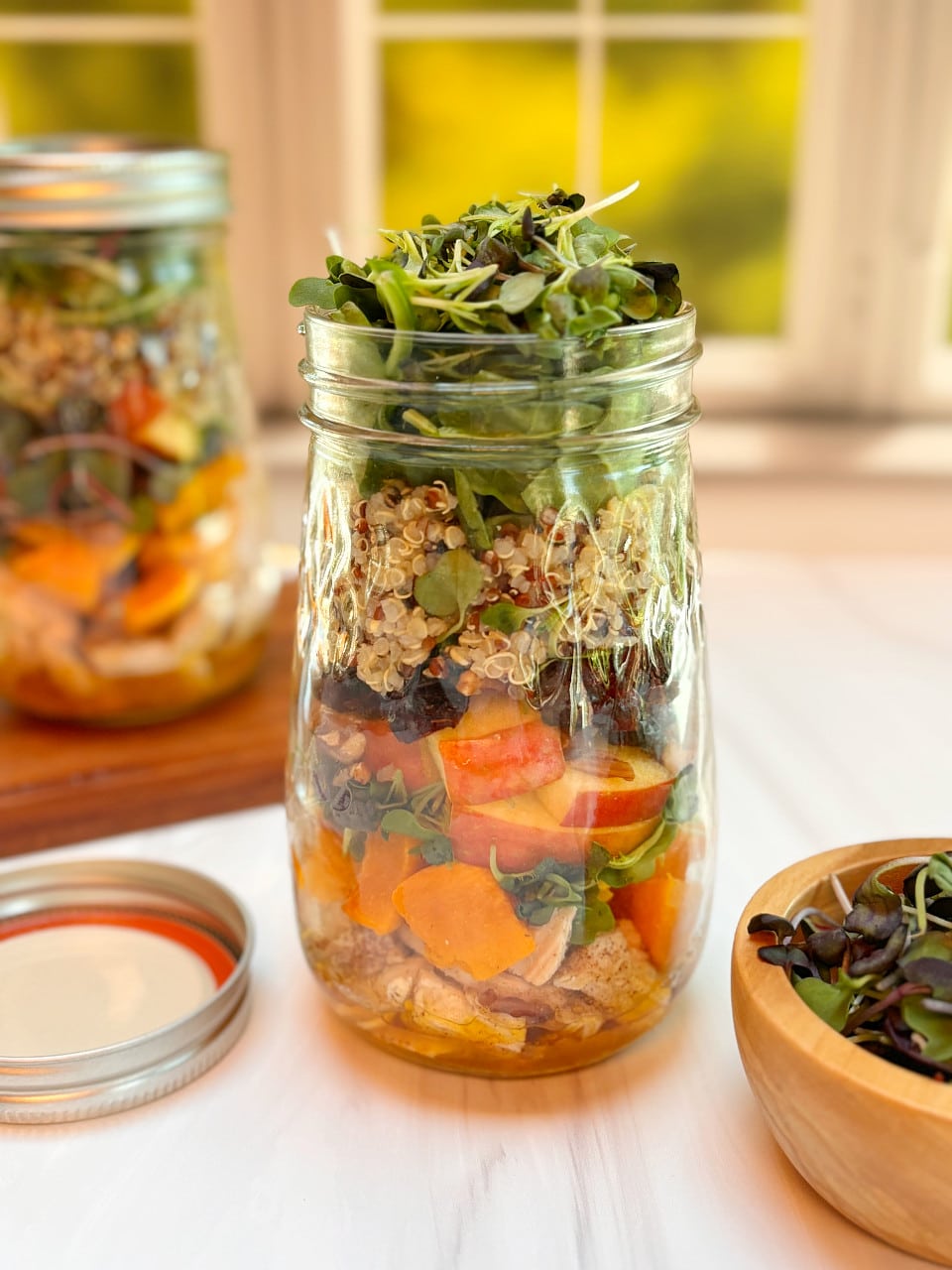 A mason jar filled with layers of roasted chicken, butternut squash, apples, quinoa and topped with rainbow microgreens