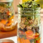 A mason jar filled with layers of roasted chicken, butternut squash, apples, quinoa and topped with microgreens on a white table. A second jar is in the background.