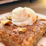 A piece of spiced apple cake with a rosette of whipped cream in the center and sprinkled with cinnamon sugar and chopped walnuts. It is on a white plate with the full cake in the background on a wire rack.