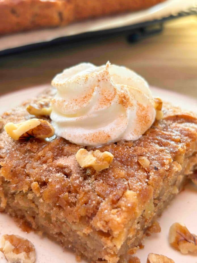 A piece of spiced apple cake with a rosette of whipped cream in the center and sprinkled with cinnamon sugar and chopped walnuts. It is on a white plate with the full cake in the background on a wire rack.