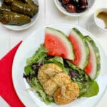 3 falafels on top of mixed greens with sliced watermelon on the side. Stuffed grape leaves and olives are in the background