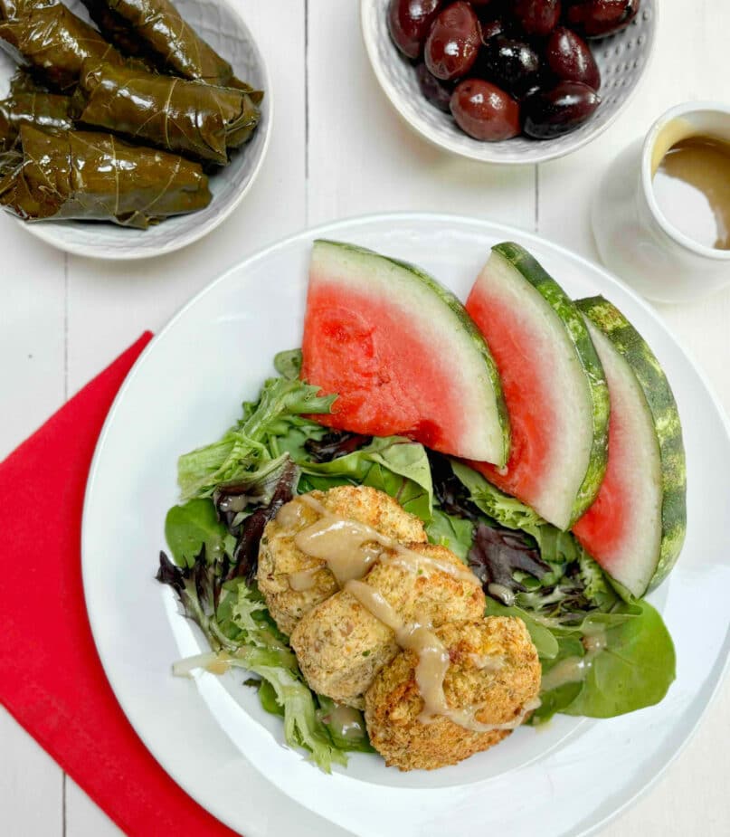 Baked falafels on a green lettuce salad with watermelon slices on the side. A dish of stuffed grape leaves and one with olives is in the background.