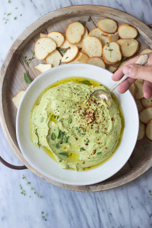 A light green dip made from beans and avocados in a white bowl with crostini crackers next to it. A hand is holding a silver spoon in the dip.