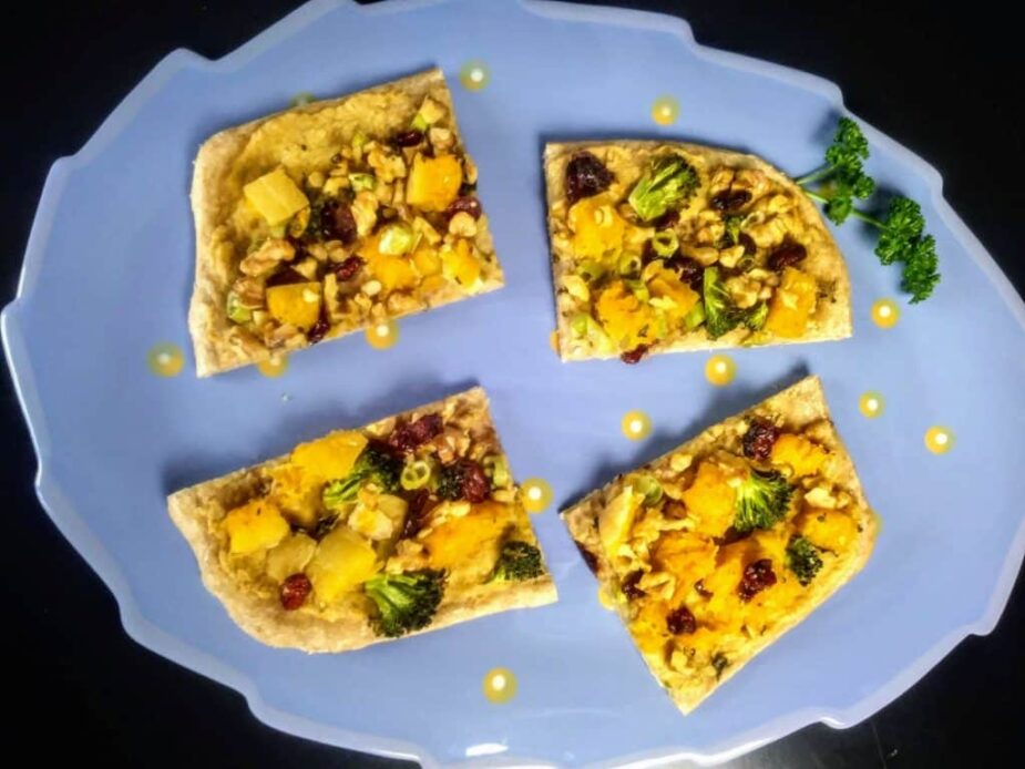 4 flatbread pieces topped with butternut squash and broccoli flowerettes and dried cranberries on a light blue platter.