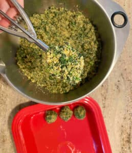 Forming the spinach mixture into balls using a large scoop with 3 formed spinach balls below in a red buttered casserole dish.