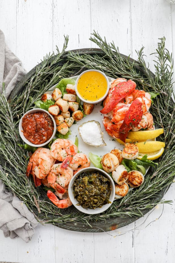 A round platter with a ring of fresh rosemary surrounding the outsied and the middle filled in with cooked shrimp, scallops, lobster and condiments making a wreath shape.