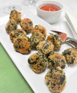 A white serving platter filled with spinach balls. A ramekin of tomato sauce and wine glasses are in the background.