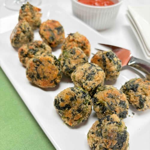 A white serving platter filled with spinach balls. A ramekin of tomato sauce and wine glasses are in the background.