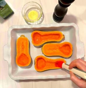 4 squash halves in a roasting pan. Someone is brushing the cut side with olive oil before roasting.