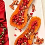 Two honeynut squash on a white platter. The squash are topped iwth pomegranate airls and candied pecans. The platter is sitting on a red napkin.