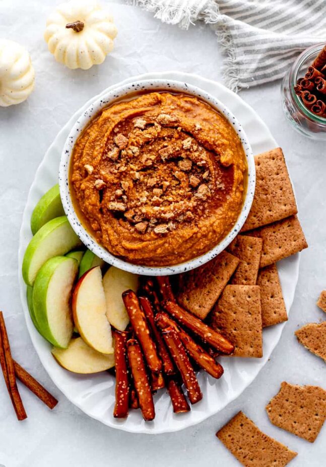 A white pottery bowl filled with pumpkin pie hummus surrounded by cut apples, whole wheat crackers and pretzel sticks on a while platter.