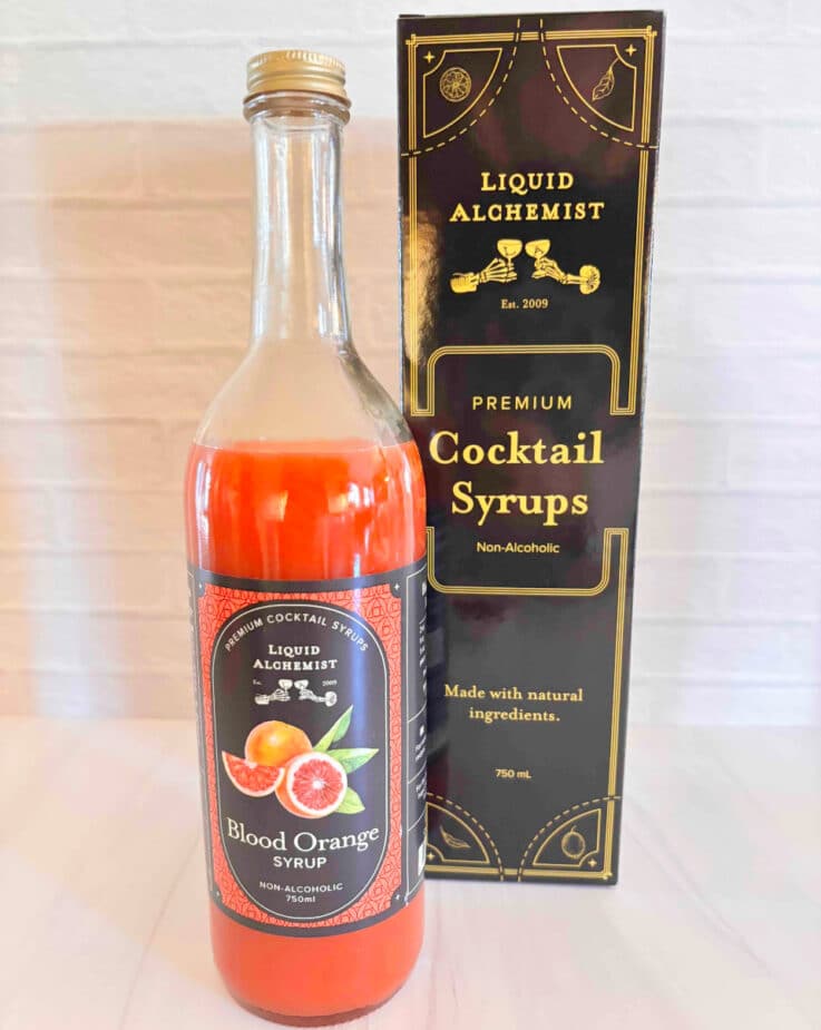 A bottle of Blood Orange Cocktail syrup by Liquid Alchemist and the box it is packaged in is sitting on a marble counter top