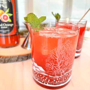 A blood orange cocktail with pomegranate juice and a sprig of mint. Another cocktail is in the background along with a bottle of blood orange cocktail syrup.