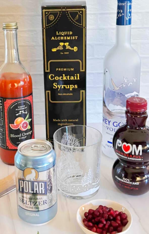 Ingredients used to make a blood orange cocktail on a marble counter top. A decorative highball cocktail glass is in the center.