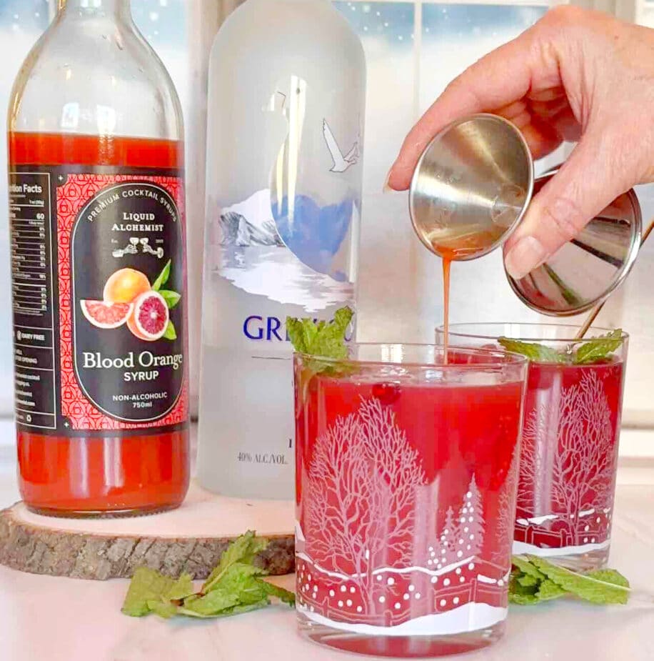 2 cocktails with a person pouring blood orange syrup into the first one. A bottle of blood orange syrup and a bottle of vodka are in the background.