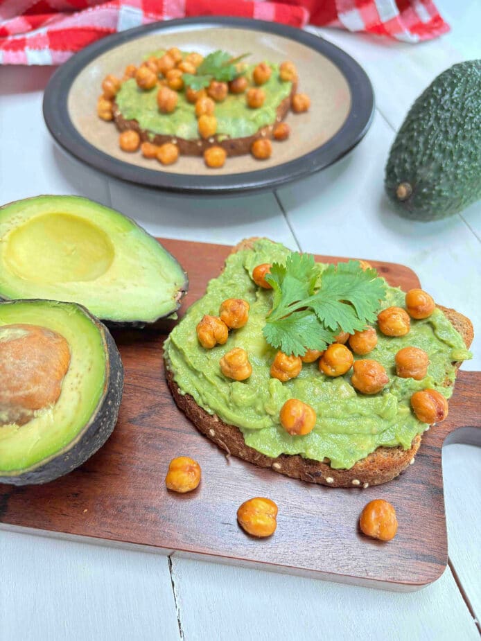 A slice of toast with smashed avocado and roasted chickpeas on top on a wooden board. Another piece is in the background.