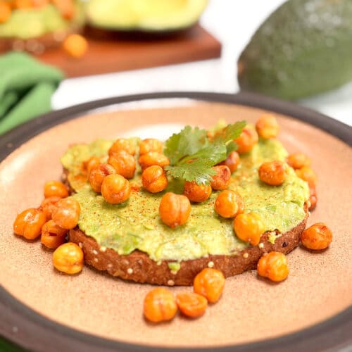 A piece of avocado toast covered with roasted chickpeas on a brown plate. A second piece and a cut avocado is in the background.