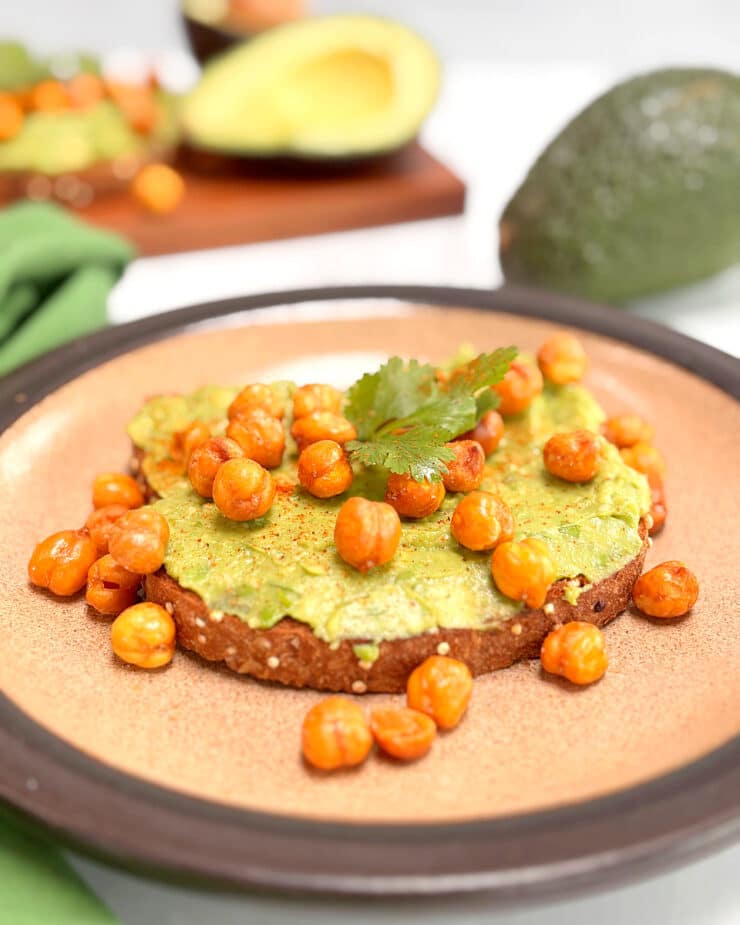 A piece of avocado toast covered with roasted chickpeas on a brown plate. A second piece and a cut avocado is in the background.