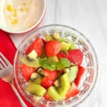 A glass bowl filled with strawberry kiwi salad with the lemon yogurt topping in the background.