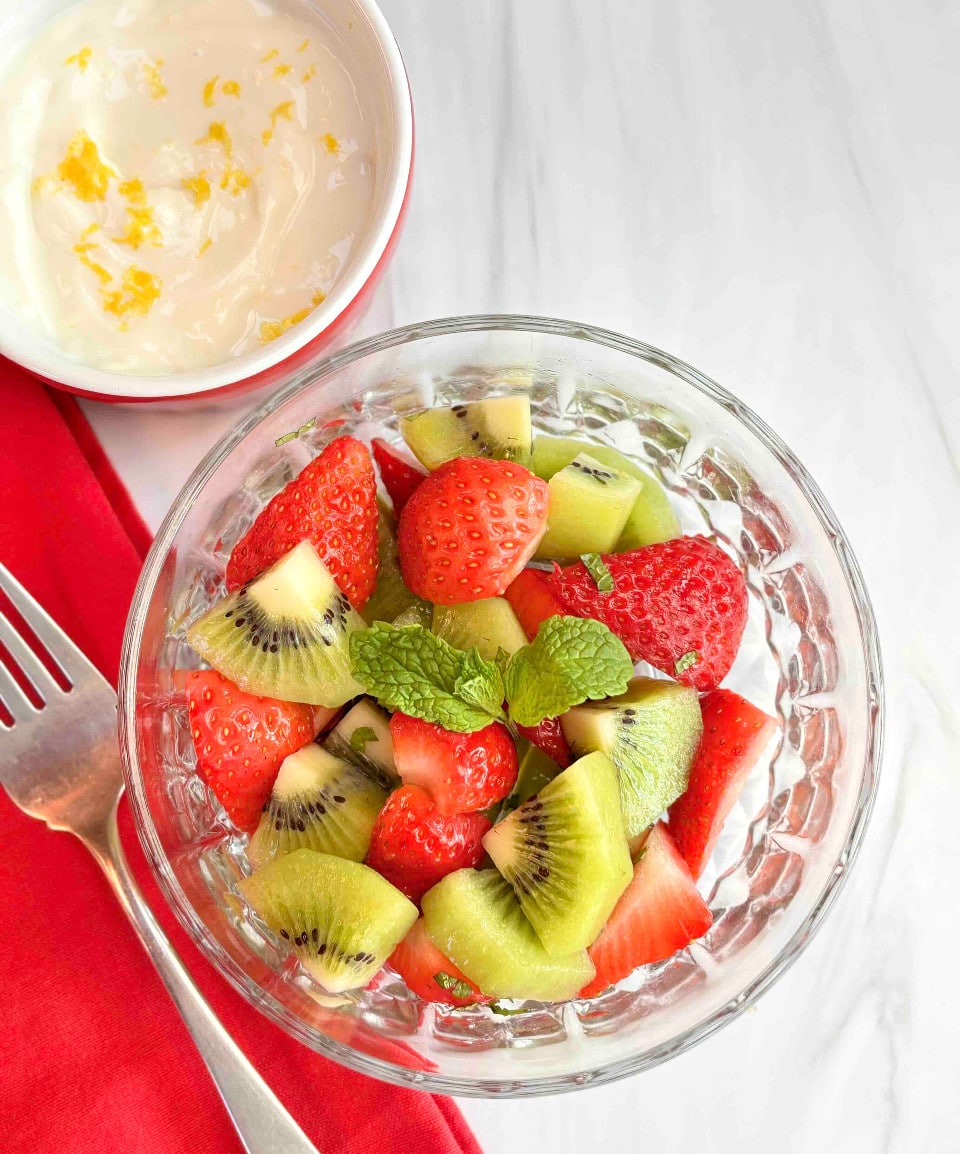 A glass bowl filled with strawberry kiwi salad with the lemon yogurt topping in the background.