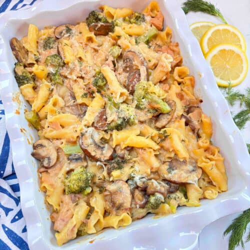 A white casserole filled with tuna casserole and a blue and white towel on the side with lemon and dill surrounding it.